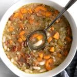 Overhead image of a white shallow bowl filled with white beans soup made with cannellini beans, carrots, onions, fresh herbs, and bacon.
