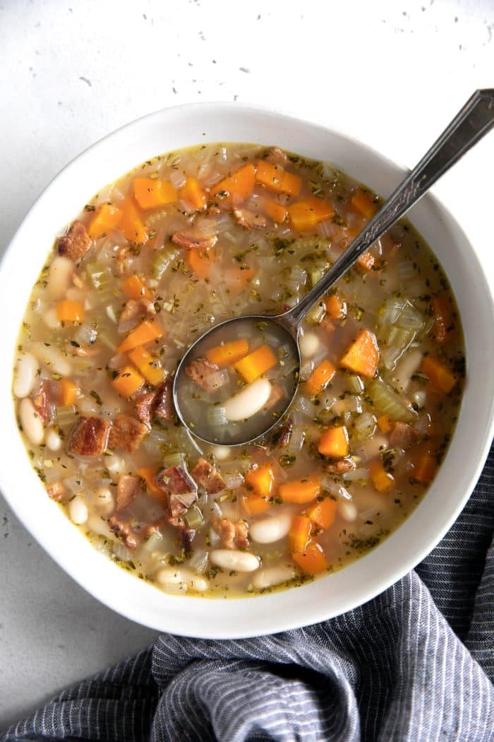 Overhead image of a white shallow bowl filled with white beans soup made with cannellini beans, carrots, onions, fresh herbs, and bacon.