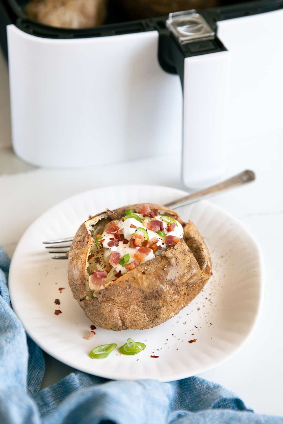 Small white plate with a baked potato topped with sour cream, bacon bits, green onions, and red pepper flakes with an air fryer basket in the background.
