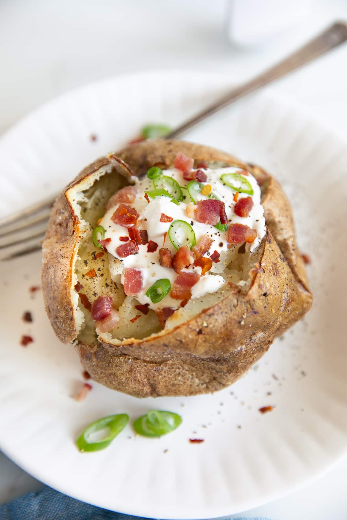 Small white plate with an air fryer baked potato sliced open and topped with sour cream, bacon bits, and green onions.