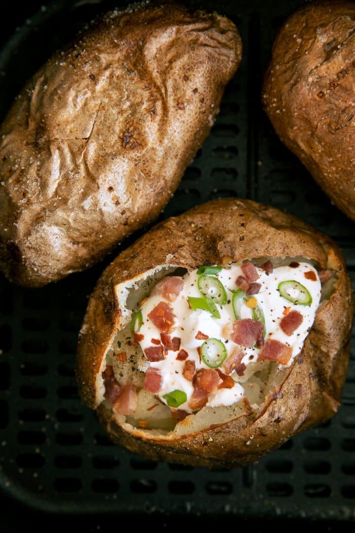 Three baked potatoes in an air fryer with one sliced open and topped with sour cream, bacon bits, and green onions.