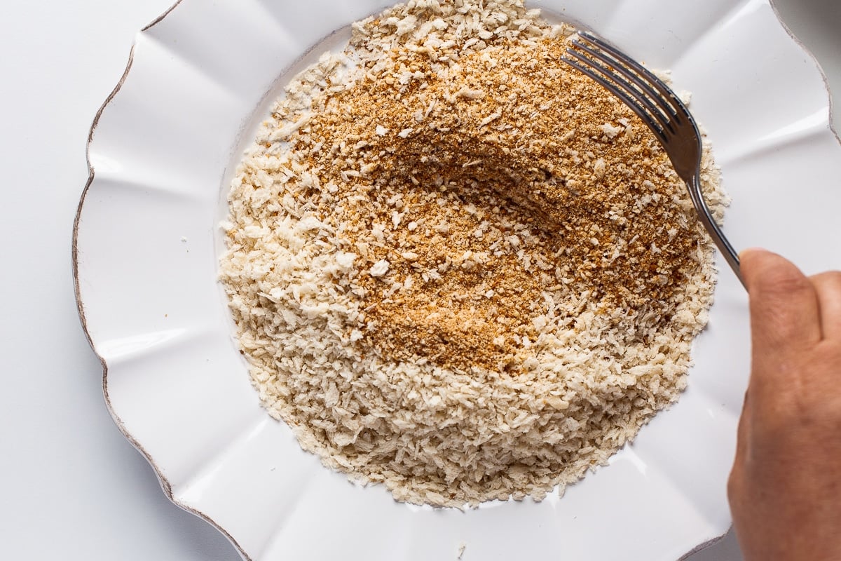 A white plate filled with a mixture of panko and plain breadcrumbs, being stirred with a fork to evenly combine for breading chicken tenders.