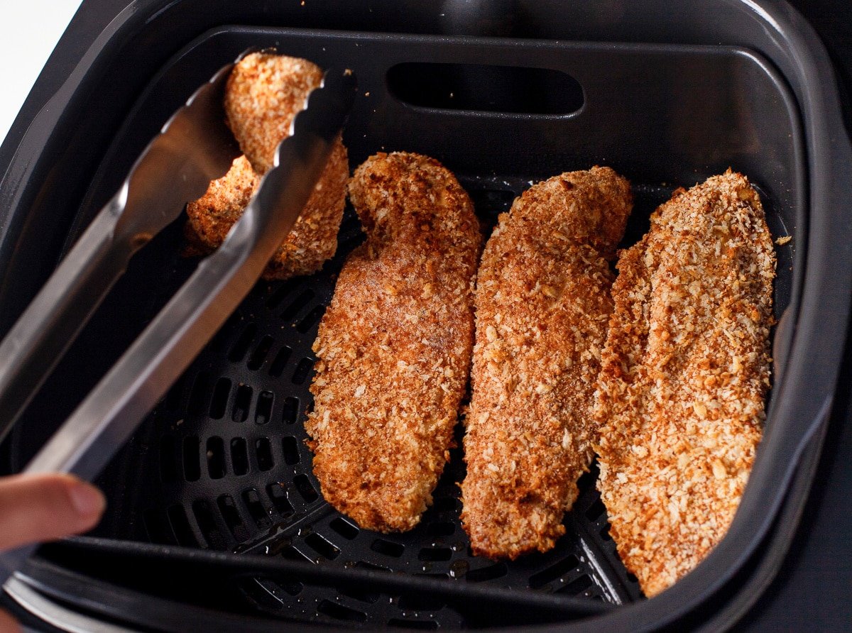 Tongs turning over breaded chicken tenders in an air fryer basket halfway through cooking at 400 degrees Fahrenheit.