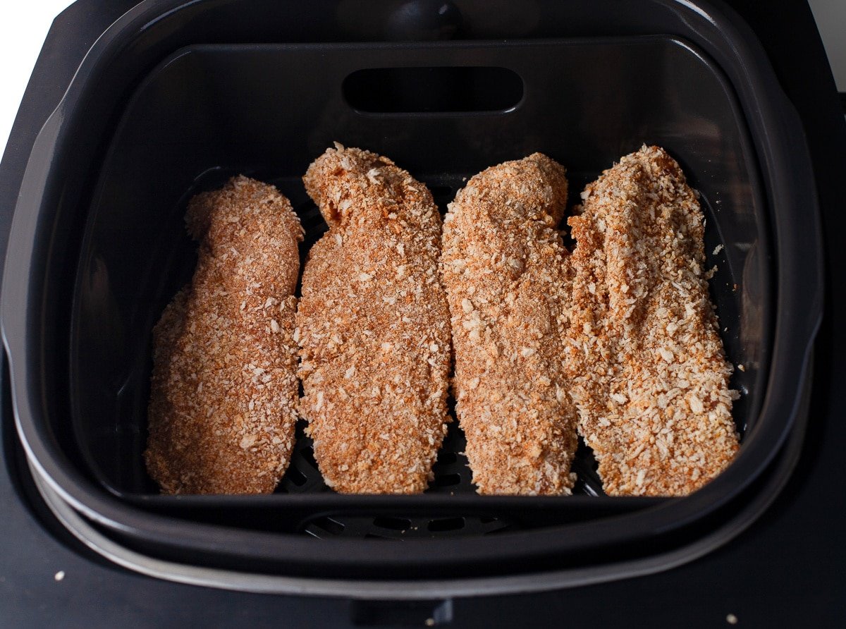 Four breaded chicken tenders arranged in a single layer inside an air fryer basket before cooking.