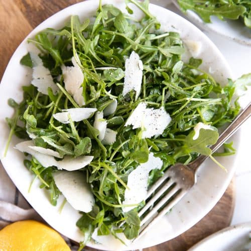 White plate with arugula and shaved parmesan salad tossed in a light lemon dressing.