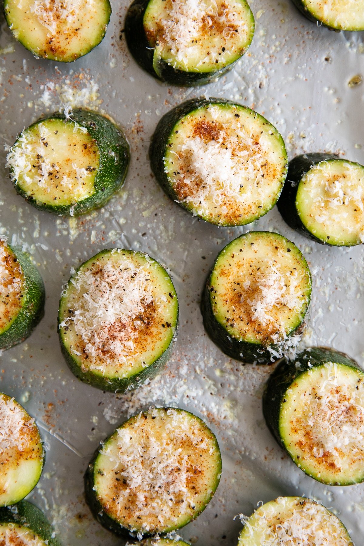 Zucchini slices on a baking sheet seasoned with salt, pepper, paprika, garlic, and topped with grated parmesan cheese.