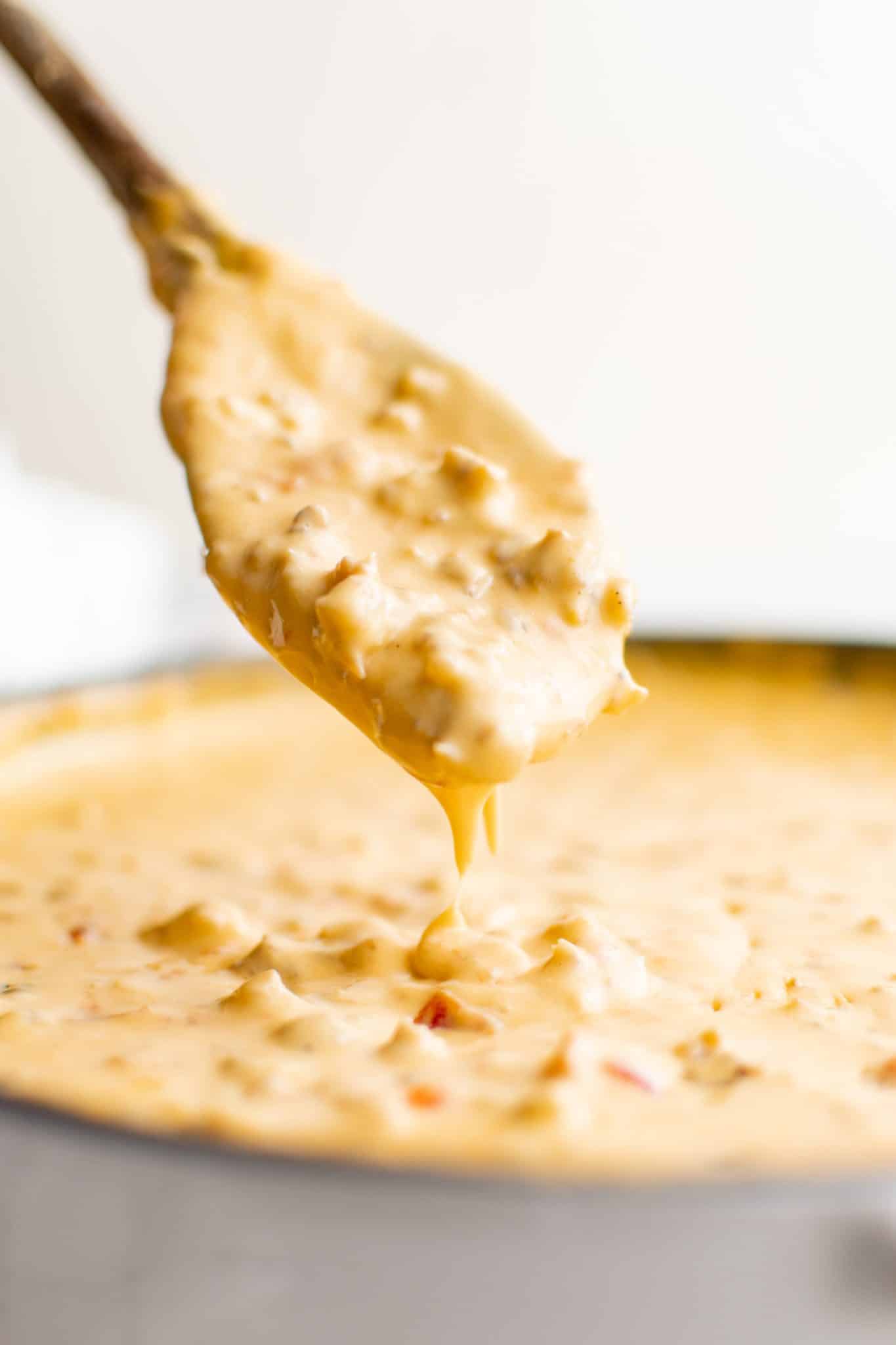 Wooden spoon lifting hot Sausage Cheese Dip with sausage and tomatoes, showing creamy texture with cheese dripping back into the skillet.