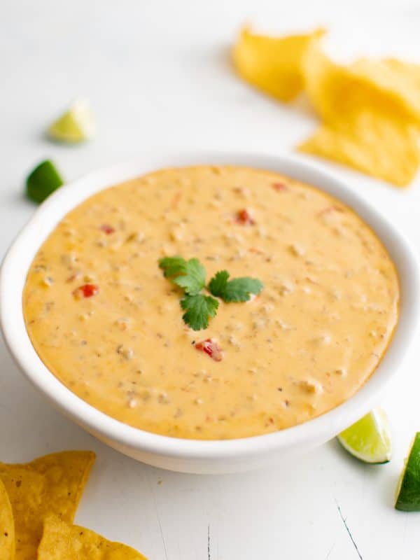 White serving bowl filled with Velveeta Cheese Dip with sausage and Rotel, topped with a cilantro sprig, surrounded by lime wedges and tortilla chips.