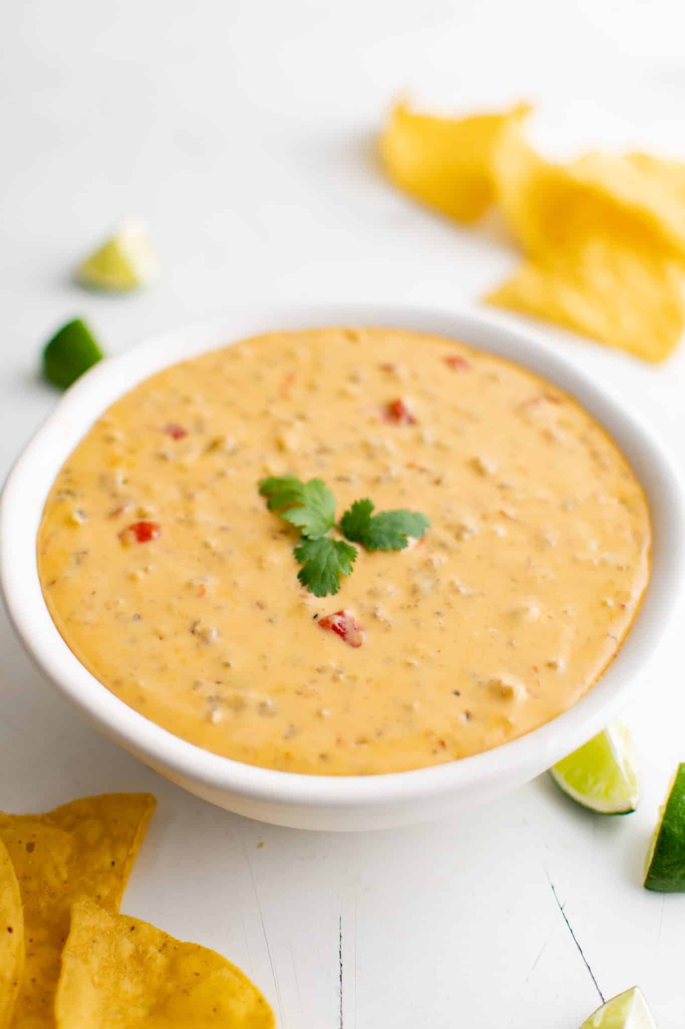 White serving bowl filled with Velveeta Cheese Dip with sausage and Rotel, topped with a cilantro sprig, surrounded by lime wedges and tortilla chips.
