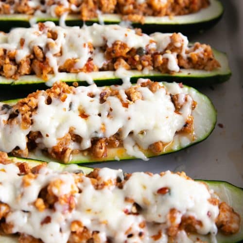 Six zucchini boats stuffed with ground sausage and topped with melted mozzarella cheese in a large white baking pan.