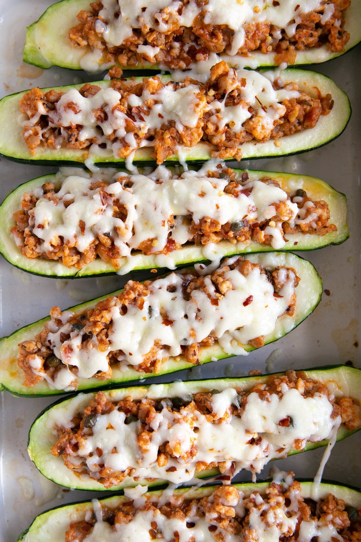 Six zucchini boats stuffed with ground sausage and topped with melted mozzarella cheese in a large white baking pan.