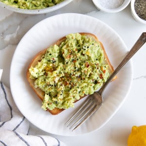 Slice of avocado toast on a small white place topped with red pepper flakes, olive oil, and black pepper.