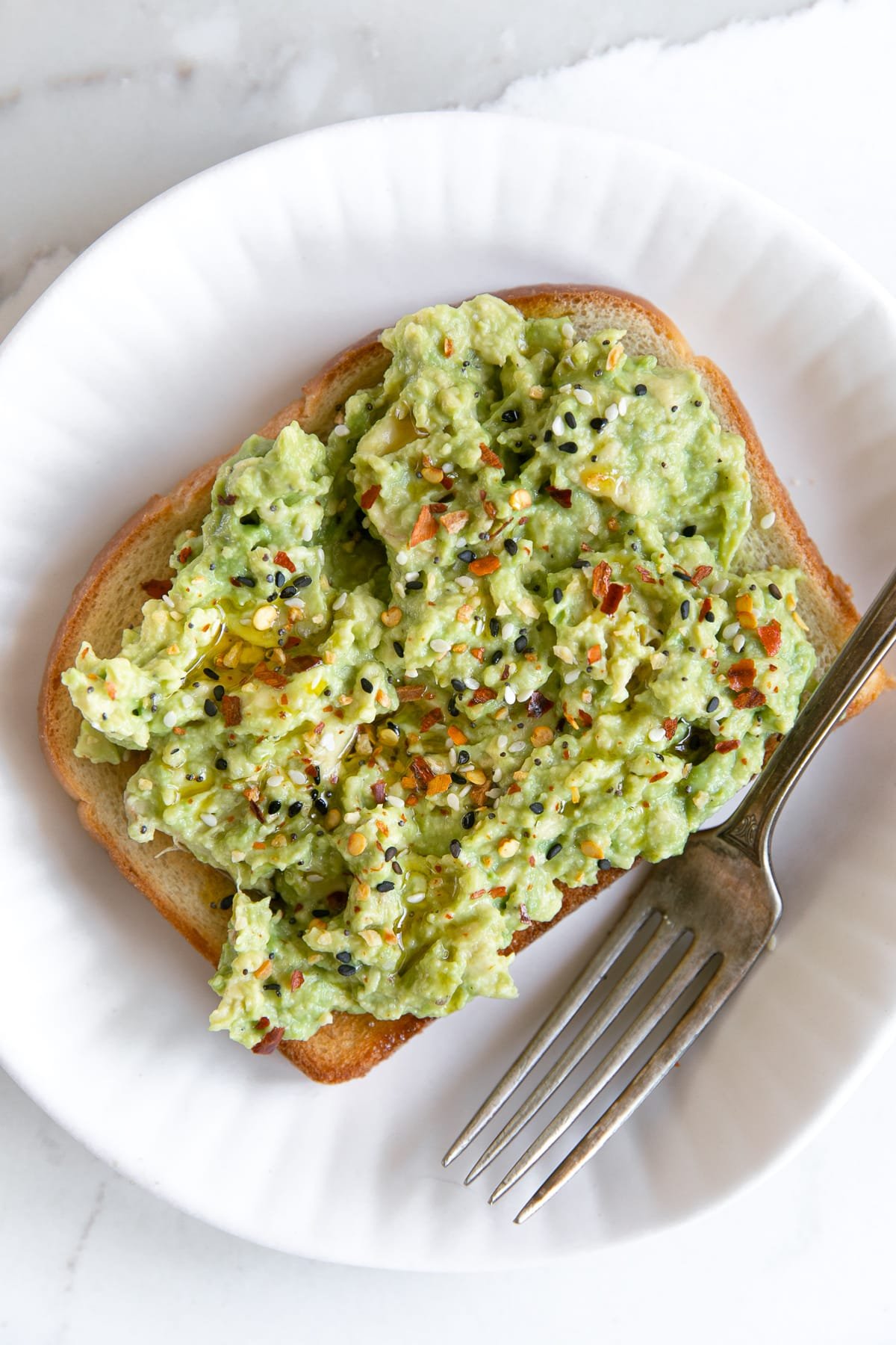 Slice of avocado toast on a small white place topped with red pepper flakes, olive oil, and black pepper.