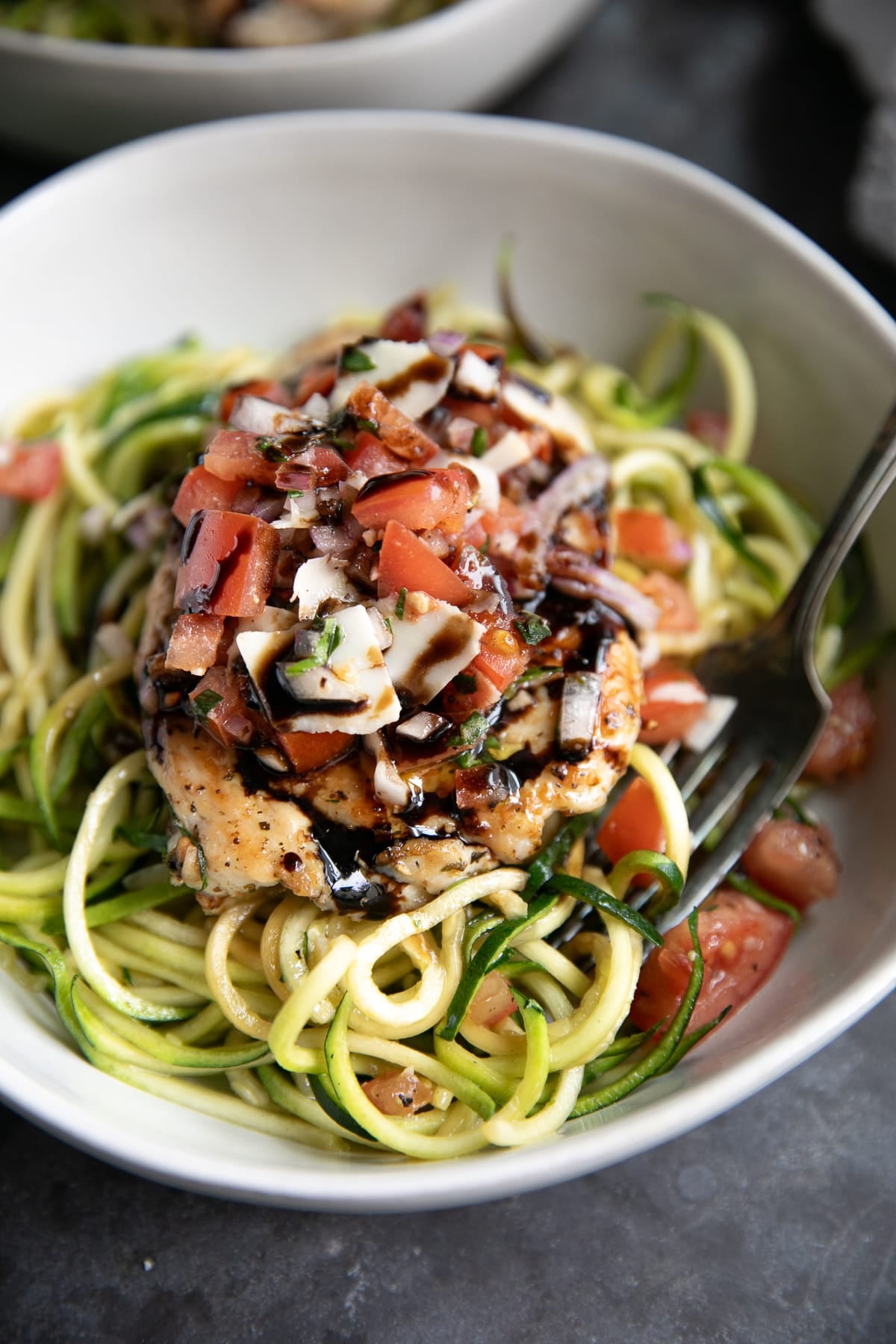 Bruschetta chicken served over green zucchini noodles in a light gray bowl, topped with chopped tomatoes, red onion, fresh mozzarella, herbs, and a dark balsamic glaze, with a silver fork along the right side.