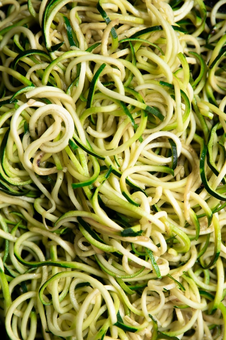 Close-up view of cooked green and pale zucchini noodles filling the entire frame, showing their long spiral shape and tender, glossy texture. No bowl, garnishes, or text overlay are visible.