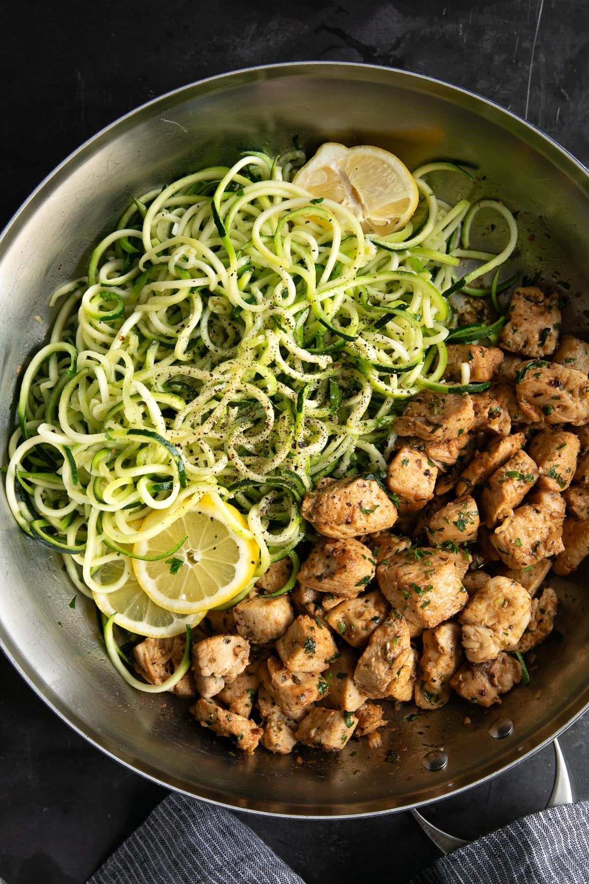 Garlic chicken bites and spiralized zucchini noodles arranged side by side in a large stainless steel skillet, with the zoodles seasoned with black pepper and garnished with fresh lemon slices and herbs.