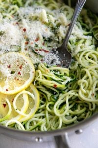 Close-up of tender green zucchini noodles in a stainless steel skillet topped with finely grated Parmesan cheese, red pepper flakes, and sliced lemon, with a silver fork resting among the noodles.