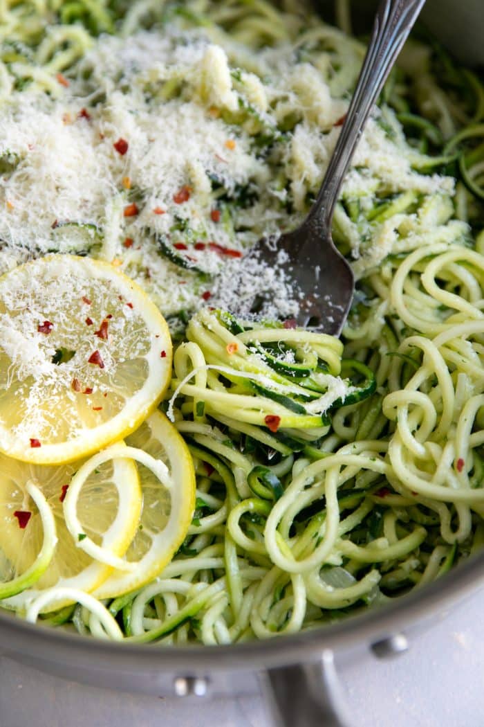 Close-up of tender green zucchini noodles in a stainless steel skillet topped with finely grated Parmesan cheese, red pepper flakes, and sliced lemon, with a silver fork resting among the noodles.