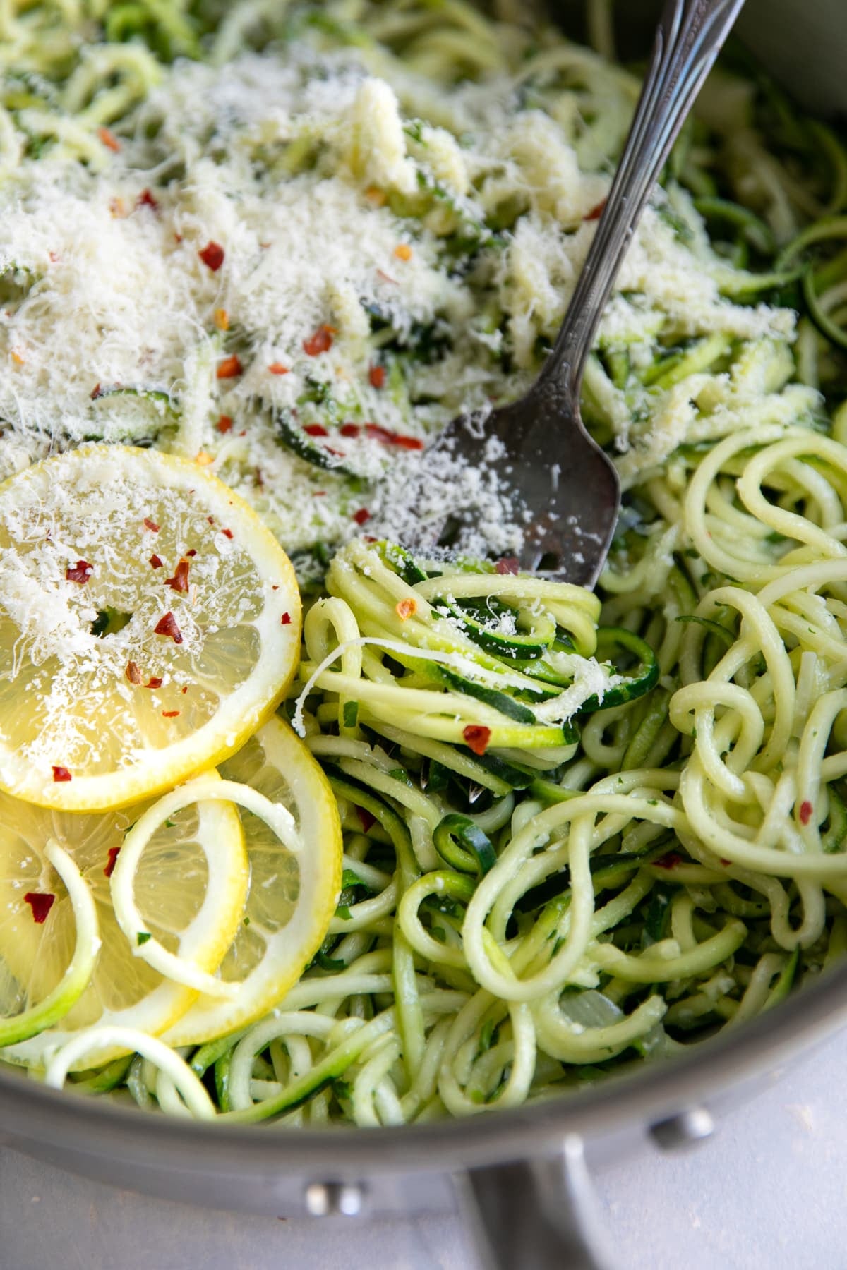 Close-up of tender green zucchini noodles in a stainless steel skillet topped with finely grated Parmesan cheese, red pepper flakes, and sliced lemon, with a silver fork resting among the noodles.