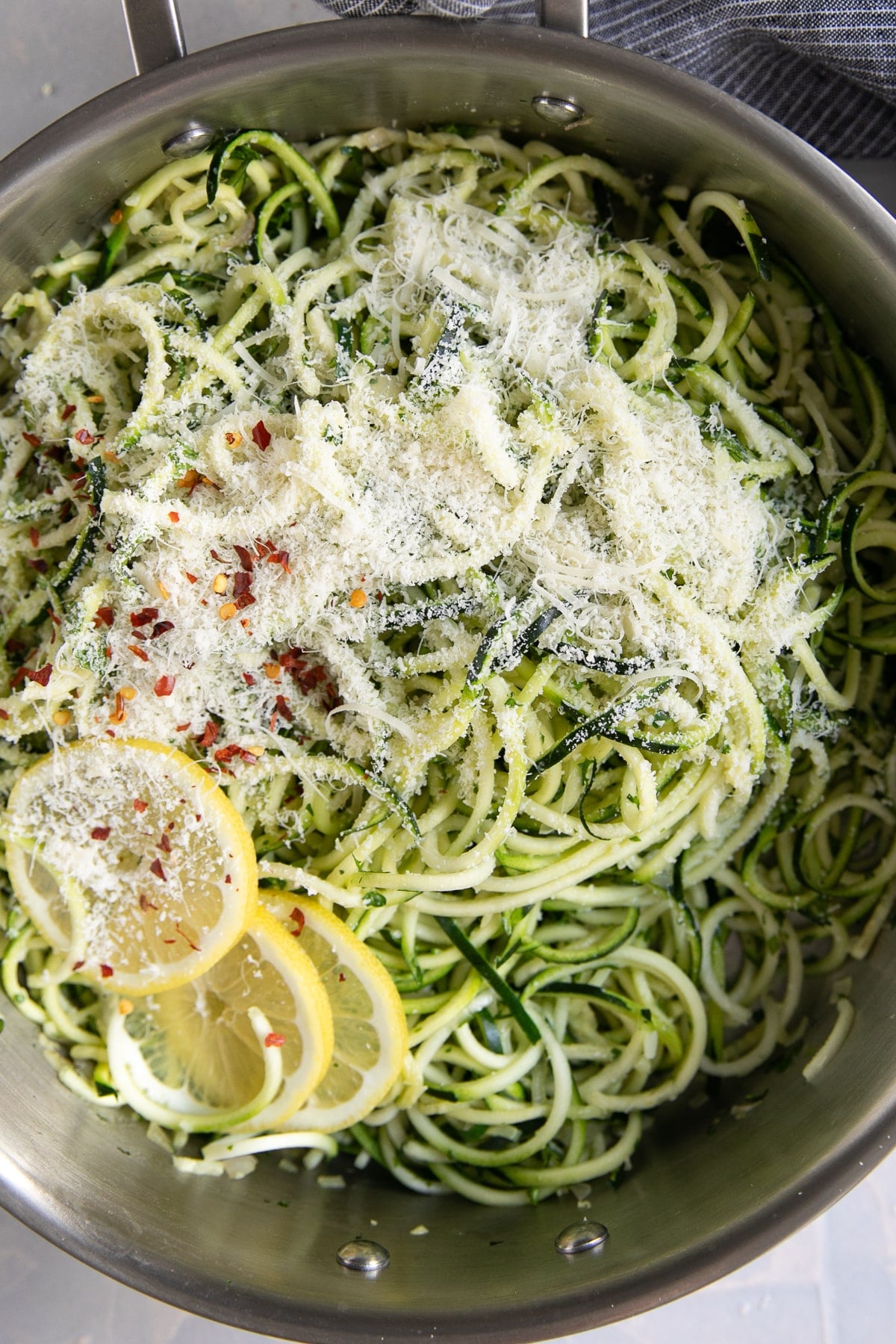 Garlic Parmesan zucchini noodles in a large stainless steel skillet topped generously with finely grated Parmesan cheese, red pepper flakes, and several fresh lemon slices. The green and pale zucchini noodles fill the pan.