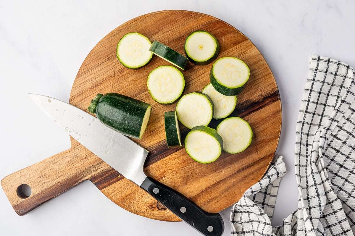 Fresh zucchini cut into thick, round slices on a large wooden cutting board. Several slices are scattered across the board with their pale centers, small seeds, and dark green skins visible. A large chef's knife rests beside the remaining piece of zucchini.