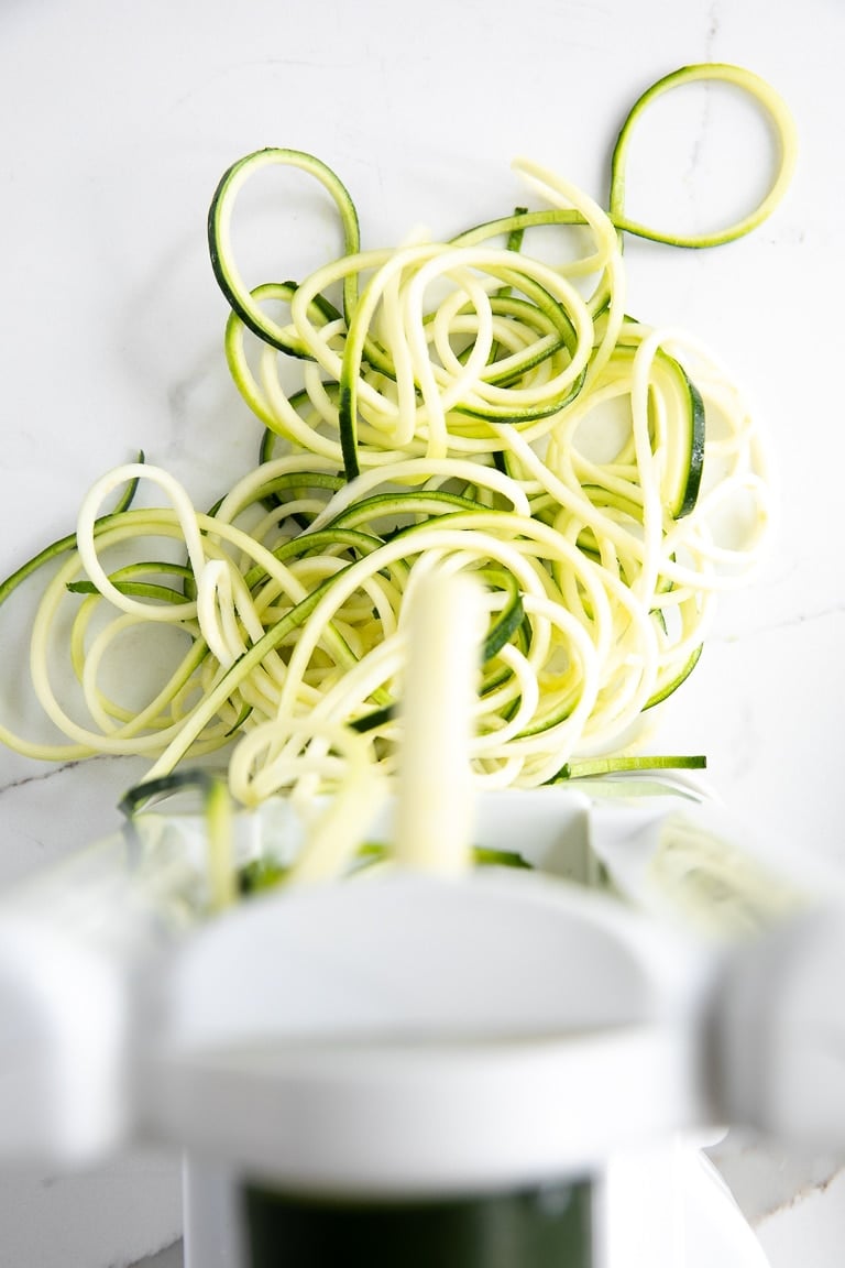 Fresh green and pale zucchini noodles coming out of a white countertop spiralizer onto a white marble surface, with long curly strands of spiralized zucchini visible behind the machine.