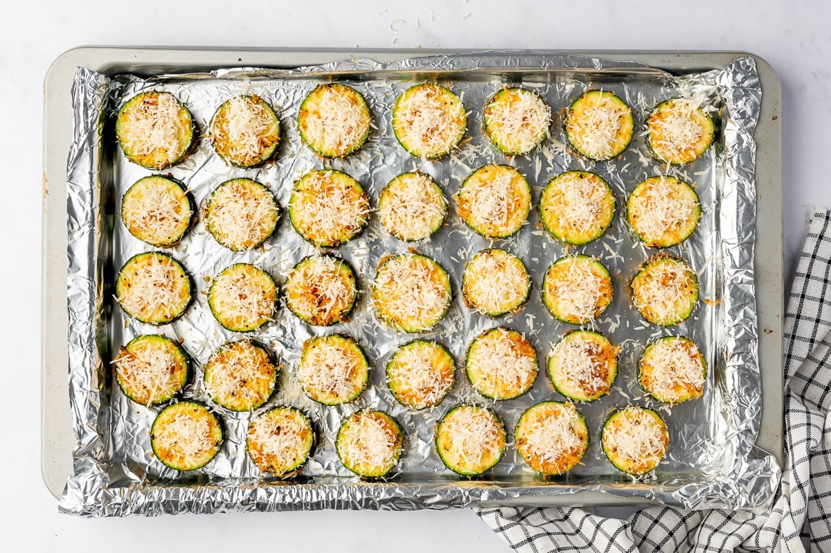 Approximately 30 thick zucchini rounds arranged in evenly spaced rows on a large aluminum foil-lined baking sheet. Each round is coated with orange-colored seasoning and topped with a generous sprinkle of finely grated Parmesan cheese before roasting.