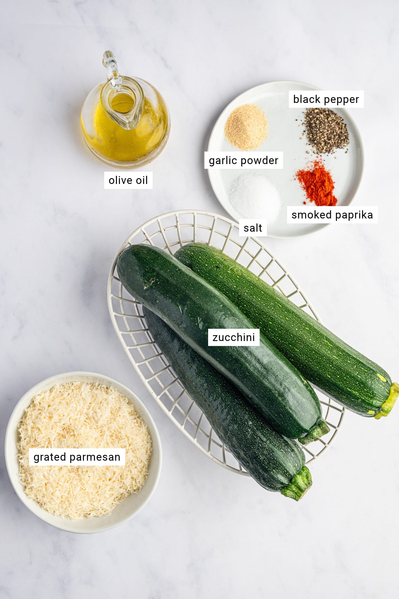 Three whole dark green zucchini in a white wire basket, surrounded by a glass bottle of olive oil, a bowl of finely grated Parmesan cheese, and small portions of garlic powder, paprika, salt, and black pepper. Each ingredient is labeled.