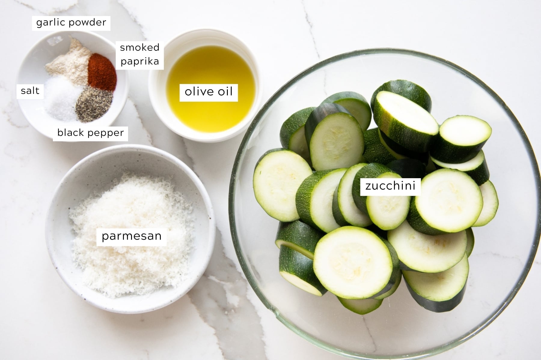 Overhead view of ingredients for roasted zucchini with Parmesan on a white marble countertop, including thick zucchini rounds in a large clear glass bowl, finely grated Parmesan cheese in a speckled light gray bowl, golden olive oil in a small white bowl, and salt, black pepper, garlic powder, and smoked paprika in a second small white bowl.