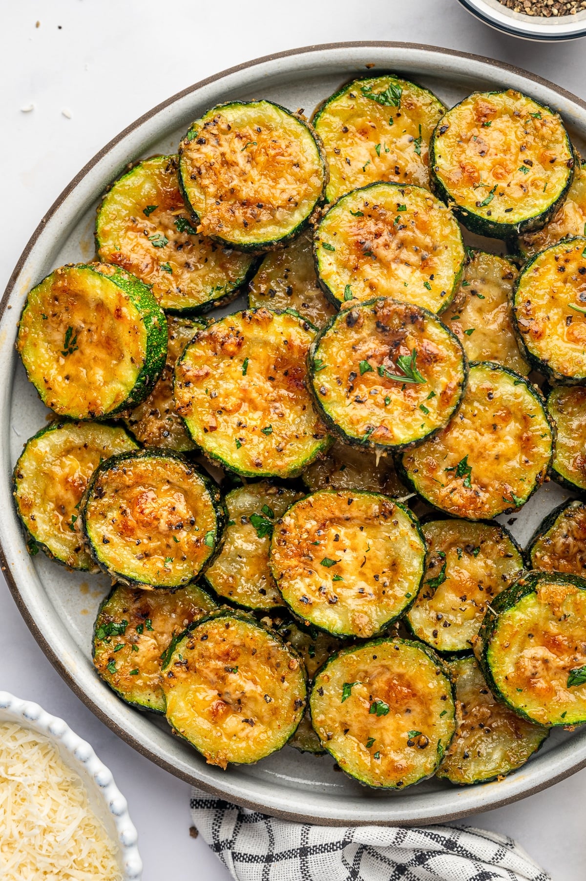 A gray ceramic plate piled high with thick roasted zucchini rounds, each topped with melted golden Parmesan cheese, black pepper, and small pieces of fresh parsley. The zucchini has dark green skins, tender yellow-green centers, and lightly browned cheese, with a few slices overlapping around the edges of the plate.