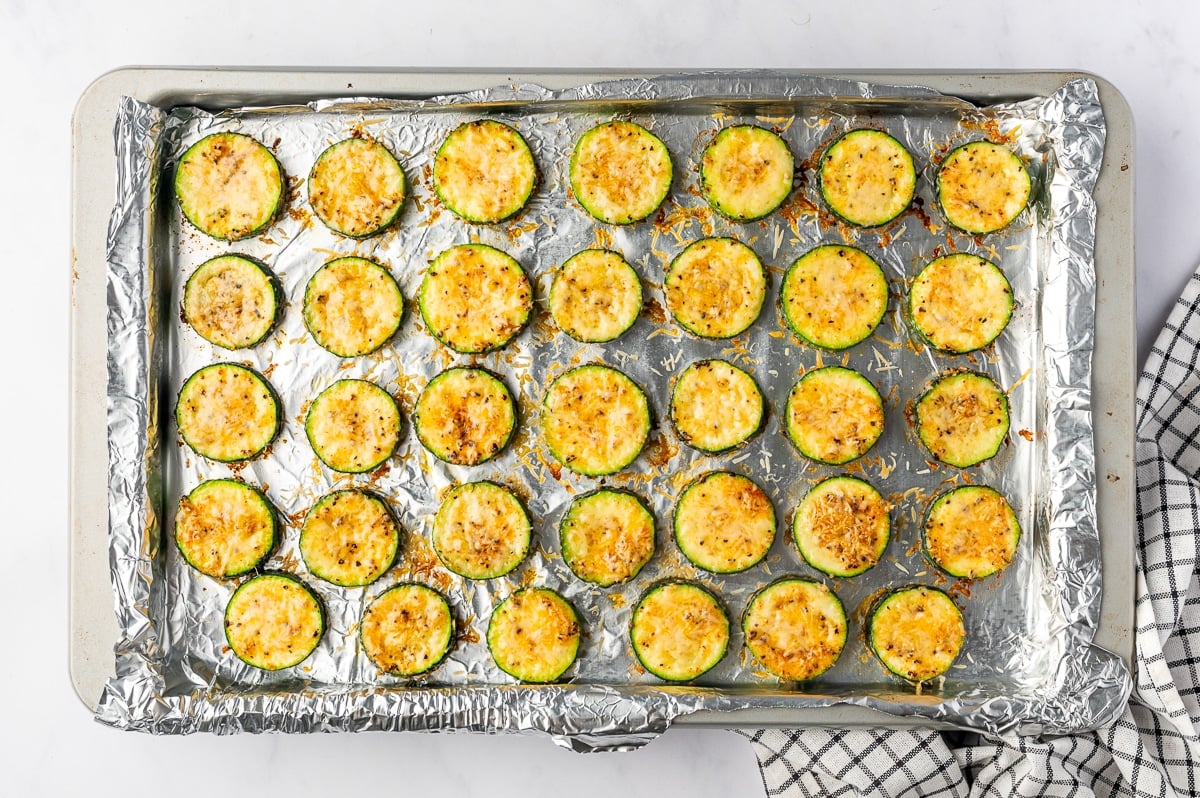 Rows of roasted zucchini rounds on a foil-lined baking sheet, with melted Parmesan cheese forming a thin golden layer over their pale yellow-green centers. Small browned spots, black pepper, and scattered pieces of toasted cheese are visible between the rounds.
