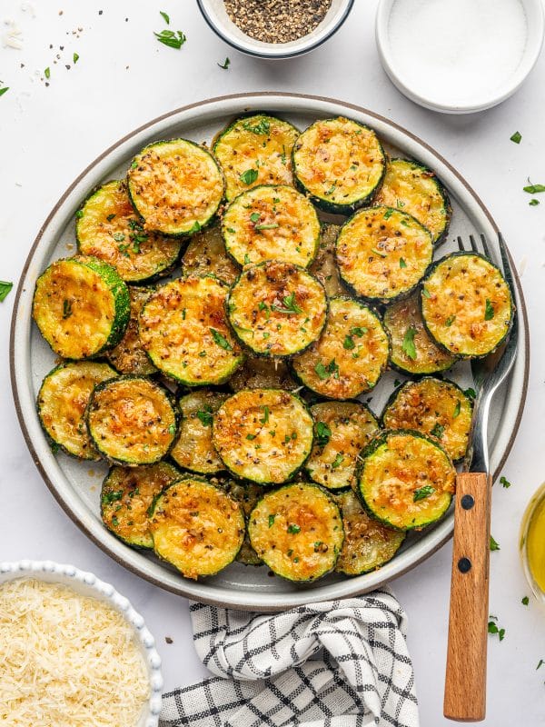 A large round gray ceramic plate filled with roasted zucchini slices topped with golden-brown Parmesan cheese, black pepper, and fresh parsley. The overlapping zucchini rounds have dark green skins and tender yellow-green centers. A wooden-handled fork rests beside the plate, with small bowls of grated Parmesan, salt, and pepper nearby and a black-and-white checked kitchen towel underneath.