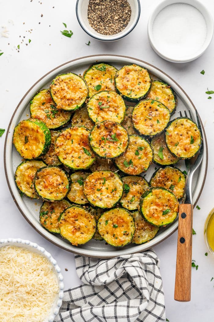 A large round gray ceramic plate filled with roasted zucchini slices topped with golden-brown Parmesan cheese, black pepper, and fresh parsley. The overlapping zucchini rounds have dark green skins and tender yellow-green centers. A wooden-handled fork rests beside the plate, with small bowls of grated Parmesan, salt, and pepper nearby and a black-and-white checked kitchen towel underneath.