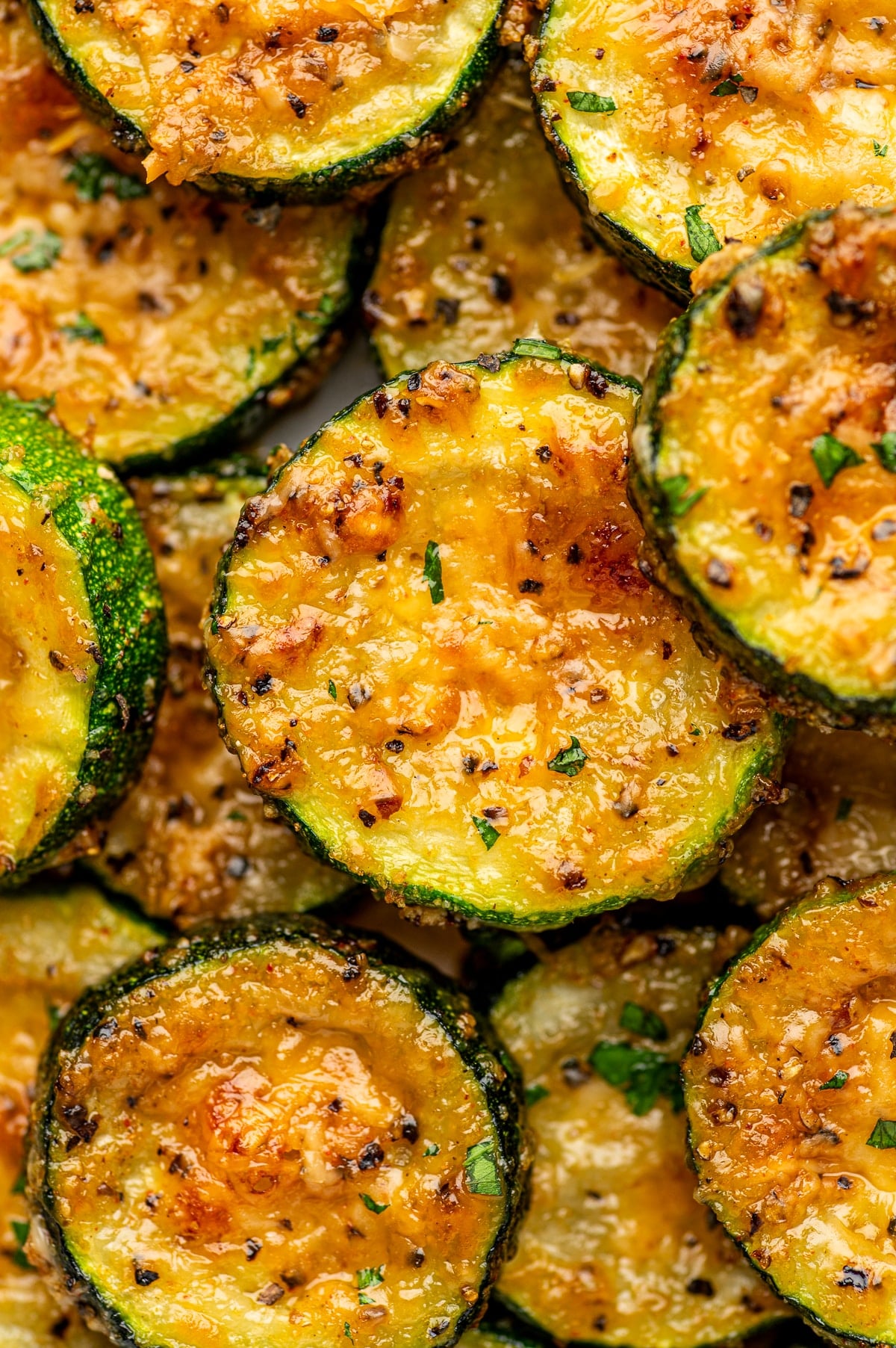 Tender roasted zucchini rounds layered together, with dark green skins and glossy yellow-green centers coated in melted Parmesan cheese. The cheese has golden-brown spots, with visible black pepper, garlic seasoning, and small pieces of chopped parsley scattered across the surfaces.