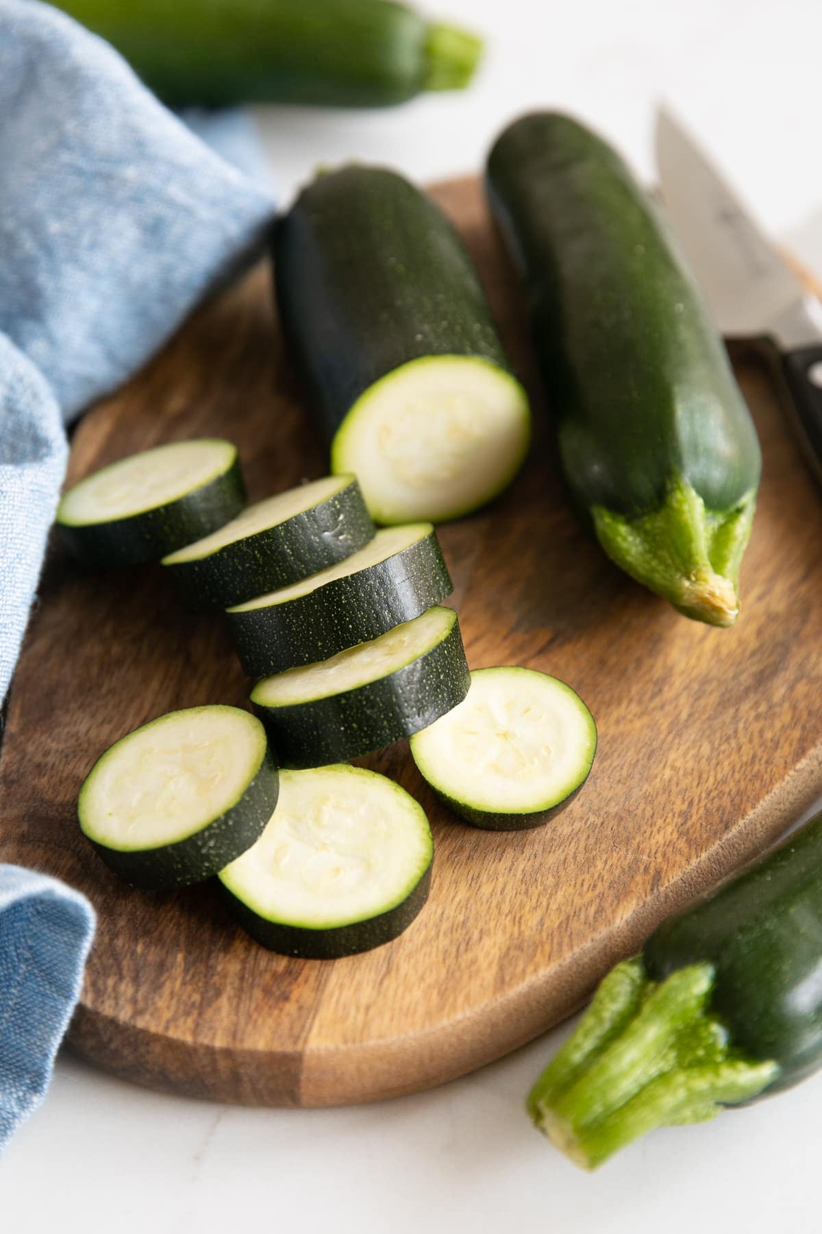Close-up of fresh dark green zucchini being cut into thick rounds on a brown wooden cutting board, with several sliced zucchini rounds in the foreground, whole zucchini in the background, a stainless steel chef's knife, and a light blue kitchen towel along the left side.