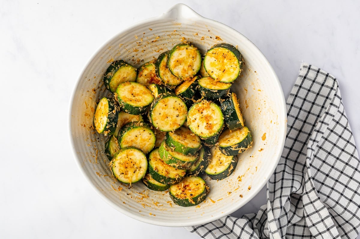 Thick zucchini rounds tossed in a large white mixing bowl, coated with olive oil, orange-red seasoning, black pepper, and finely grated Parmesan cheese. The seasonings cling to the pale zucchini centers and dark green skins, with some collecting along the inside of the bowl.