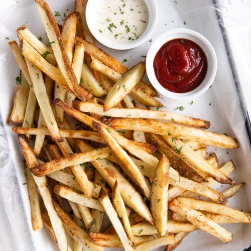White tray filled with french fries cooked in the air fryer and garnished with chopped parsley and served with a side of ketchup and ranch dressing.