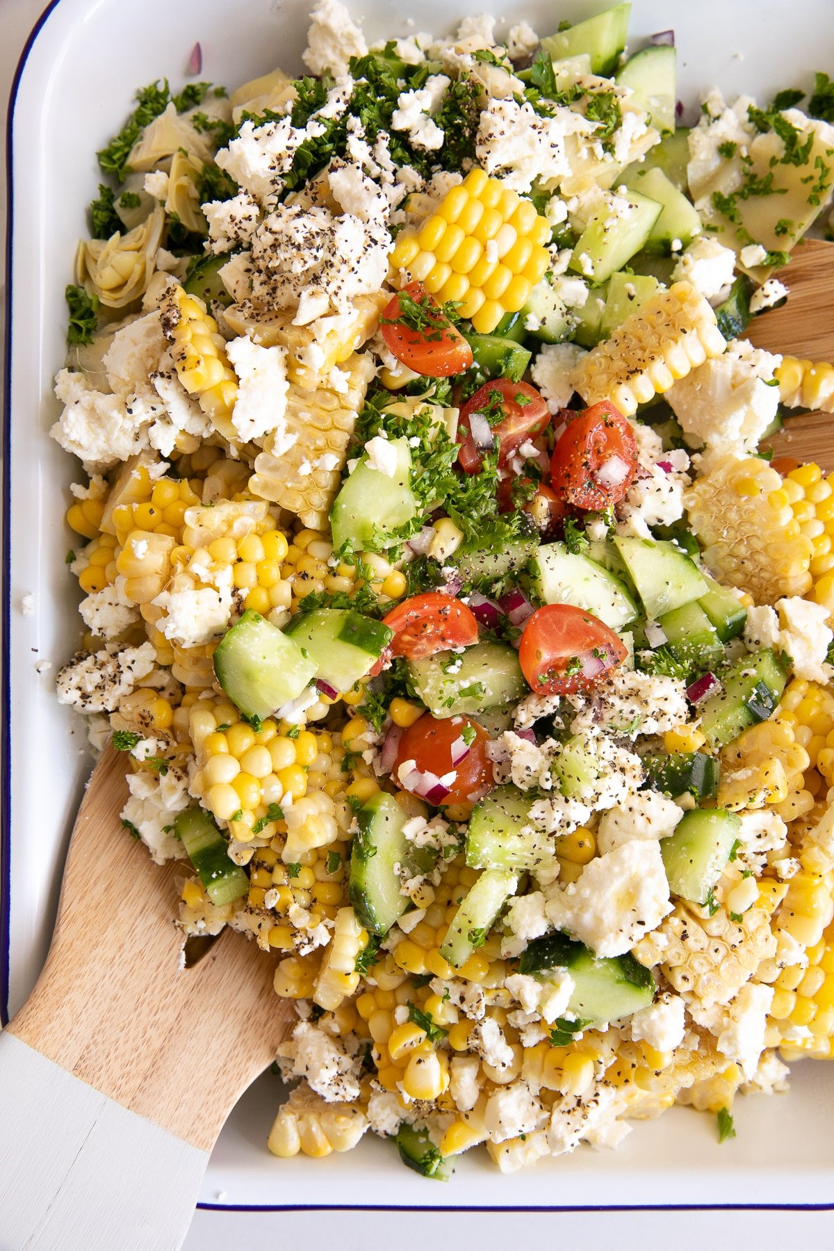 Platter filled with all of the ingredients for corn salad including corn, cucumber, feta cheese, artichoke hearts, and cherry tomatoes.