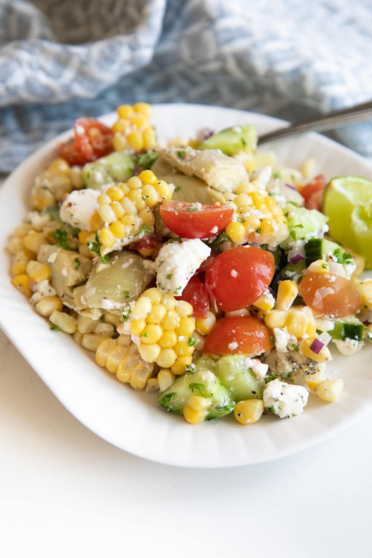 Small serving plate filled with fresh corn salad made with corn, cherry tomatoes, cucumber, feta cheese, and artichoke hearts.