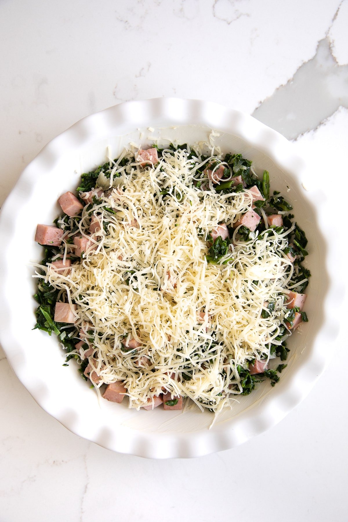 White pie pan filled with cooked spinach, shallots, cubes of smoked ham, and shredded cheese.