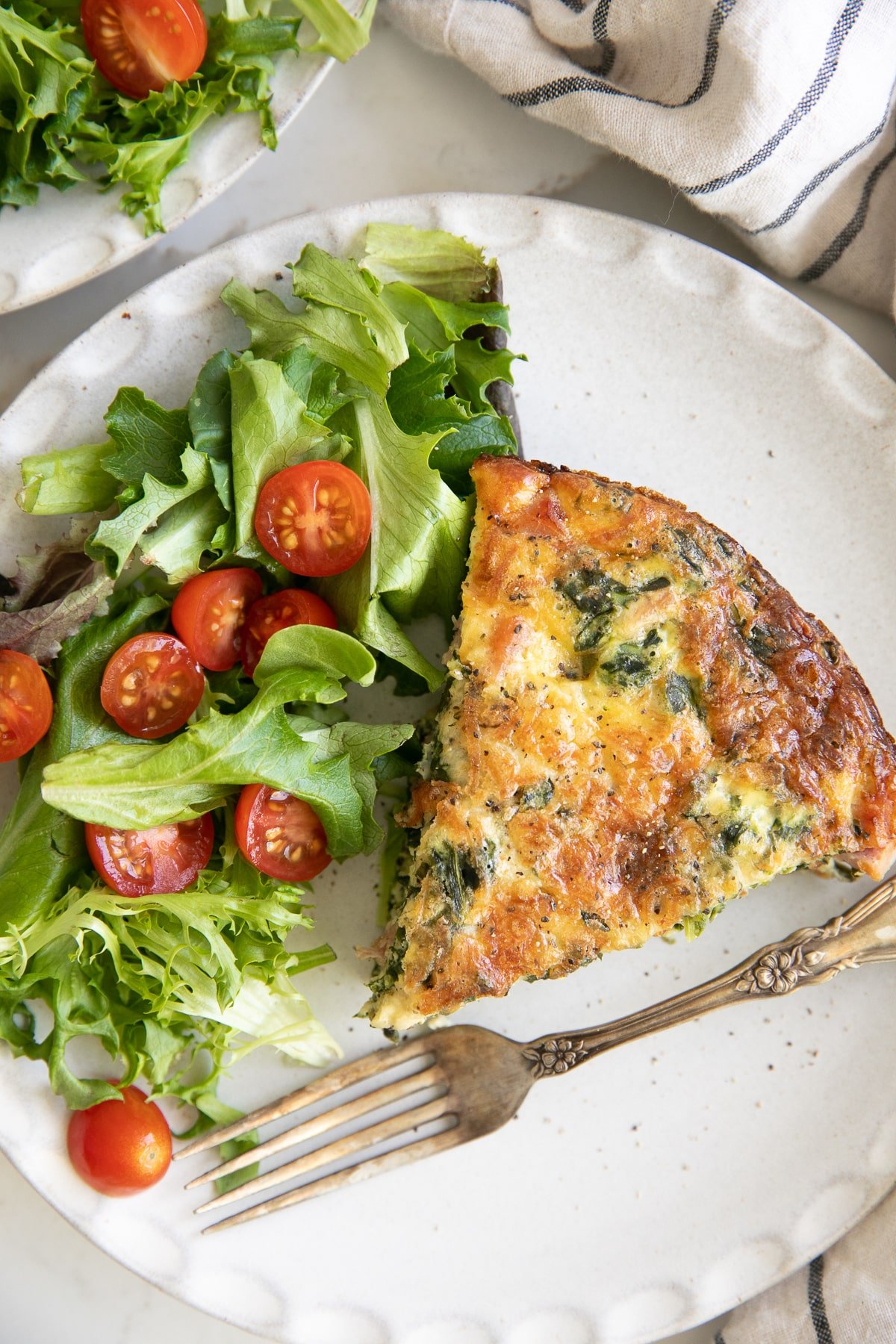 White plate with a large slice of crustless quiche with spinach and ham and a side of spring salad with tomatoes.
