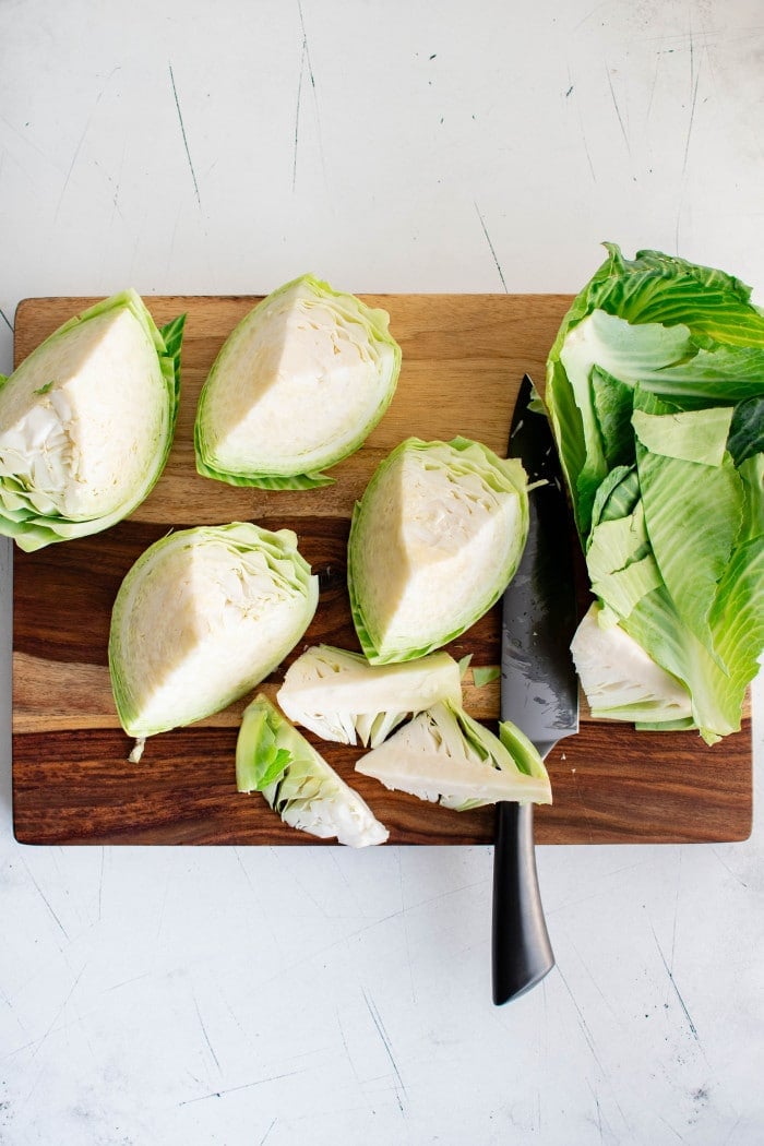 A large head of green cabbage cut into wedges with the outer leaves removed, shown on a wooden cutting board with a black-handled chef’s knife.