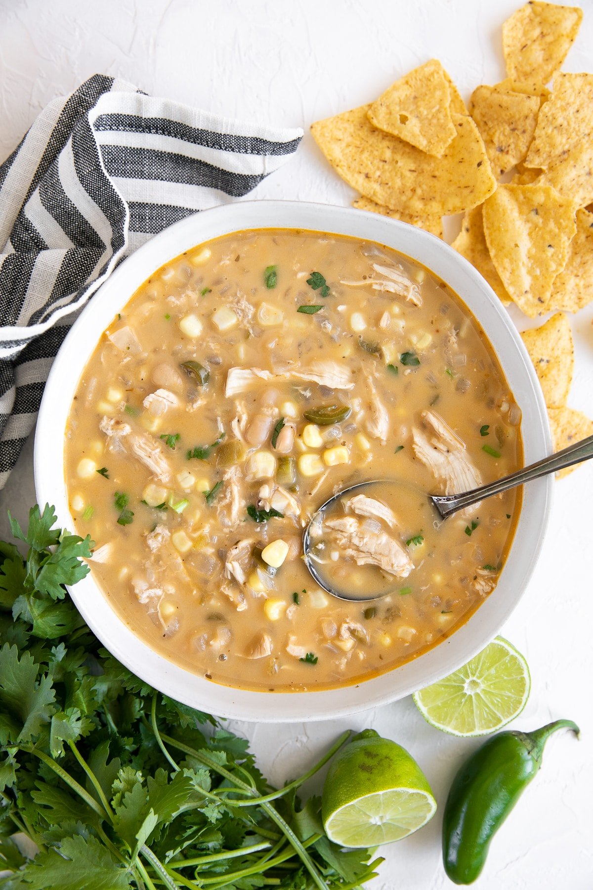 White shallow soup bowl surrounded by fresh cilantro, limes, and tortilla chips filled with creamy white chicken chili garnished wit crushed corn tortilla chips and chopped cilantro.