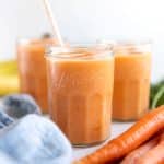 Three glasses filled with icy cold carrot smoothie with fresh carrots and a light blue cloth napkin set beside.