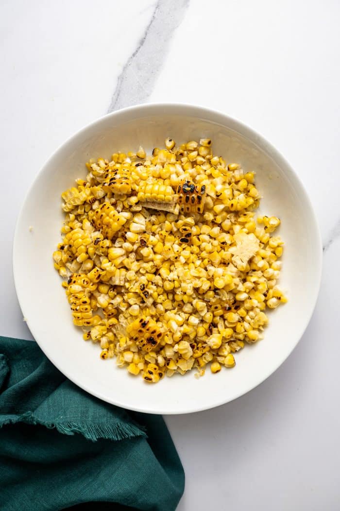 Overhead image of grilled corn kernels cut from the cob and piled on a large round white plate. The kernels are bright yellow with scattered blackened charred bits and a few small cut rounds of corn still mixed in. A dark teal cloth napkin sits in the lower left corner on the white marble background.