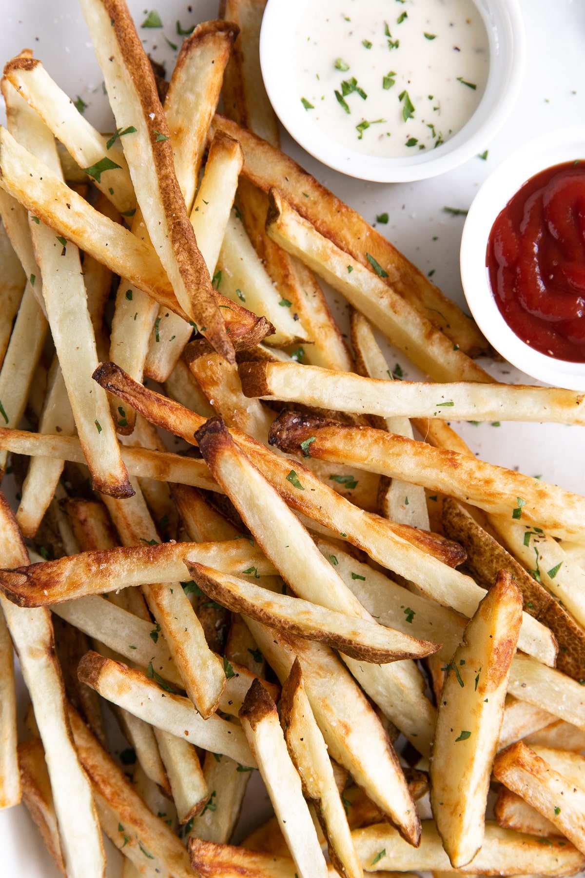 Close-up shot of crispy air fryer French fries topped with parsley, with ketchup and ranch dressing in the background.