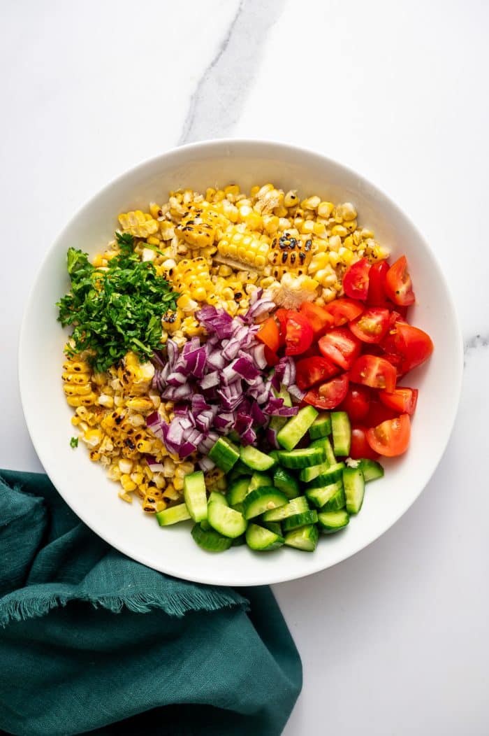 Overhead image of a large white bowl filled with the corn salad ingredients before mixing. Charred grilled corn kernels fill the bowl and are topped with chopped fresh cilantro, diced red onion, chopped cucumber, and halved cherry tomatoes. A dark teal cloth napkin sits beside the bowl on the white marble surface.