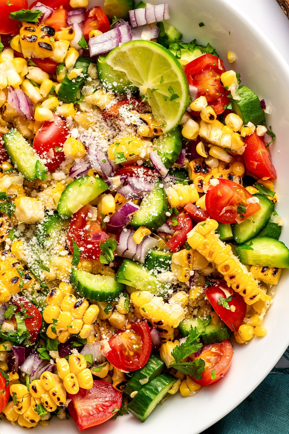 Close-up image of corn salad inside a white bowl. Visible ingredients include charred yellow corn kernels, chopped cucumber, halved red cherry tomatoes, diced red onion, chopped cilantro, crumbled Cotija cheese, and a lime wedge resting on top. The textures of the vegetables and grill marks on the corn are clearly visible.