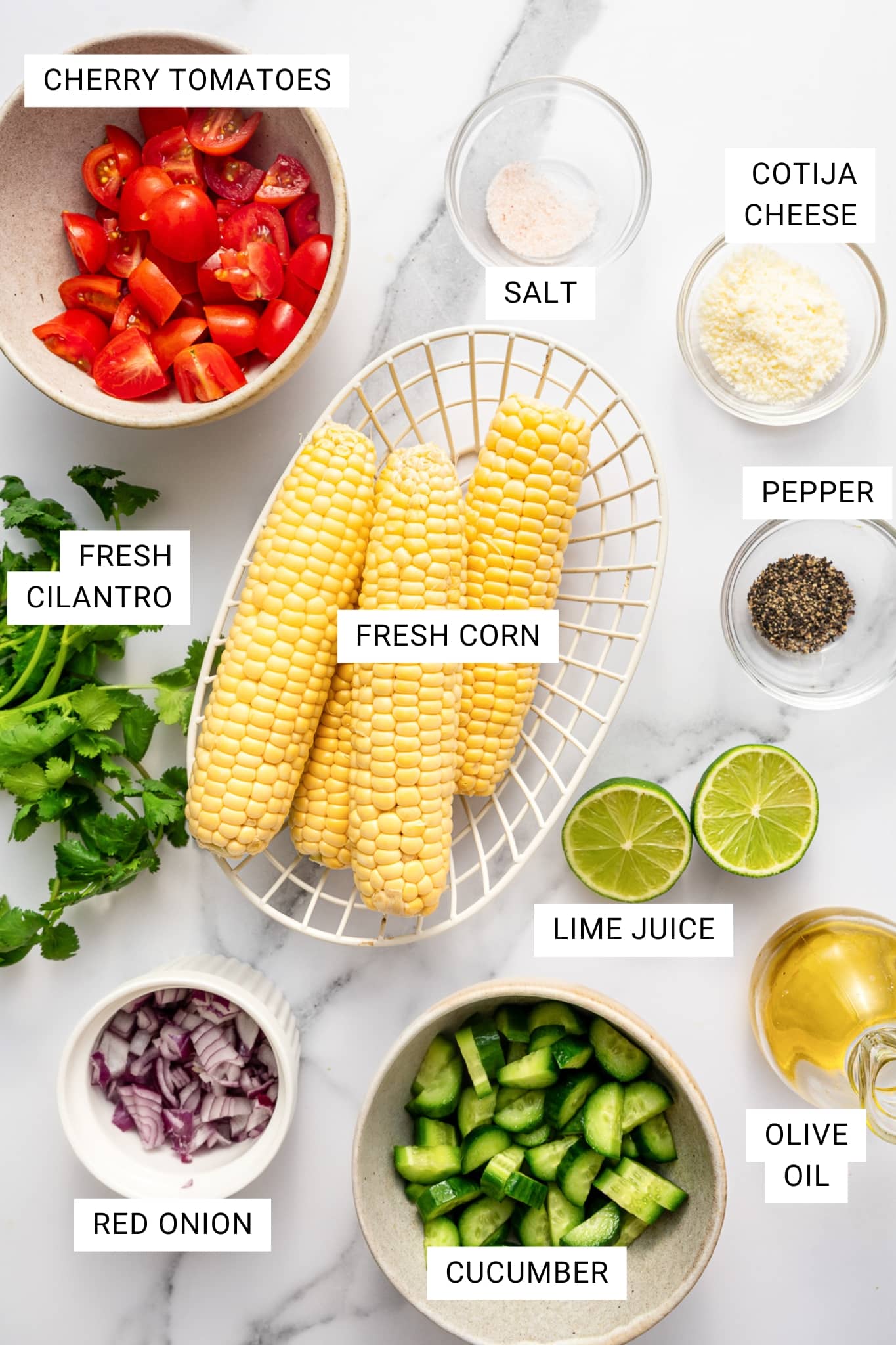 Overhead image of the ingredients for corn salad arranged on a white marble surface with text overlays labeling each ingredient. Four ears of fresh corn sit in a white wire basket in the center. Surrounding them are a beige bowl of halved red cherry tomatoes, fresh cilantro, a small white ramekin of diced red onion, a beige bowl of chopped cucumber, halved limes labeled lime juice, a glass container of olive oil, a small glass bowl of salt, a small glass bowl of black pepper, and a small glass bowl of crumbled Cotija cheese.