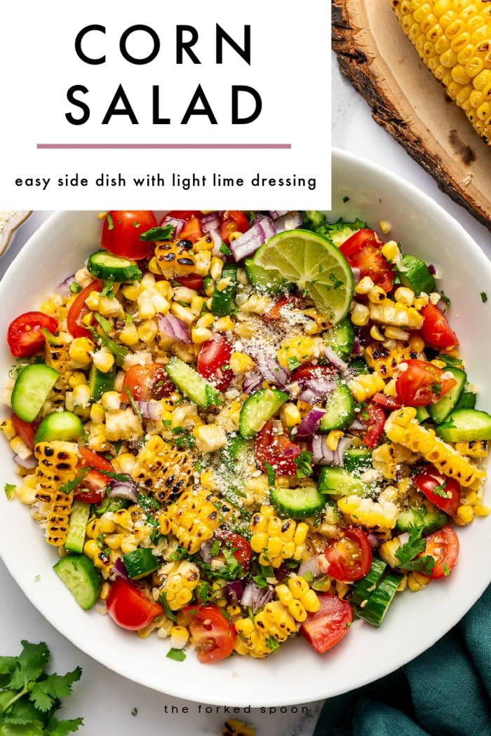 Pinterest pin for corn salad featuring a large overhead photo of corn salad in a white bowl on a white marble surface. The salad is made with charred yellow corn, chopped cucumber, halved cherry tomatoes, diced red onion, chopped cilantro, a sprinkle of Cotija cheese, and a lime wedge garnish. A grilled ear of corn on a round wooden board is partially visible in the top right corner, and a dark teal cloth napkin appears along the lower right edge. A large light gray text box overlays the upper left portion of the image with bold black text that reads “CORN SALAD,” a thin muted pink line underneath, and smaller black text that reads “easy side dish with light lime dressing.” The words “the forked spoon” appear at the bottom.